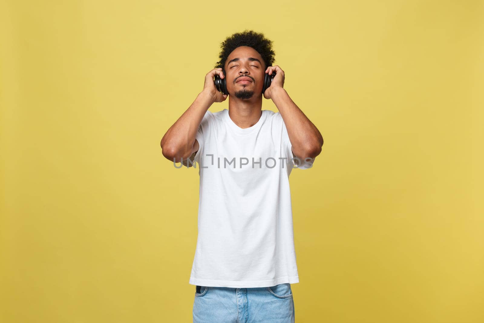 Close up portrait of dark skinned man with closed eyes, has headphones, listens to favourite music. Teenager enjoys audio track isolated on yellow background