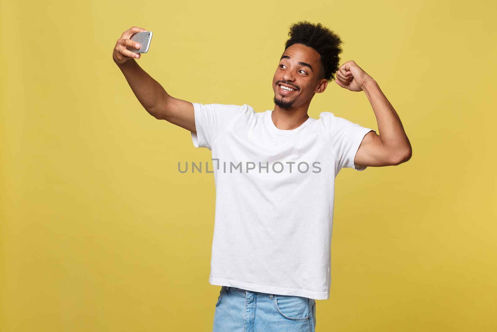 young afro american black man smiling happy taking selfie self portrait picture with mobile phone looking excited having fun posing cool isolated in yellow background in communication technology.