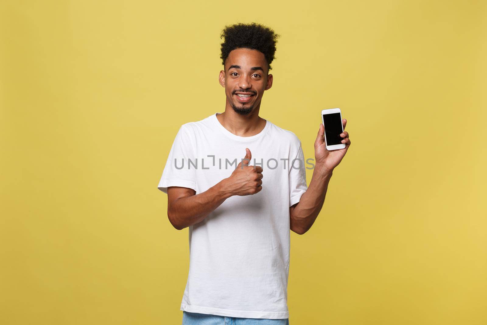 Portrait of attractive young black man isolated on yellow background, holding blank smartphone, smiling at camera, showing thubms up gesture, feeling happy