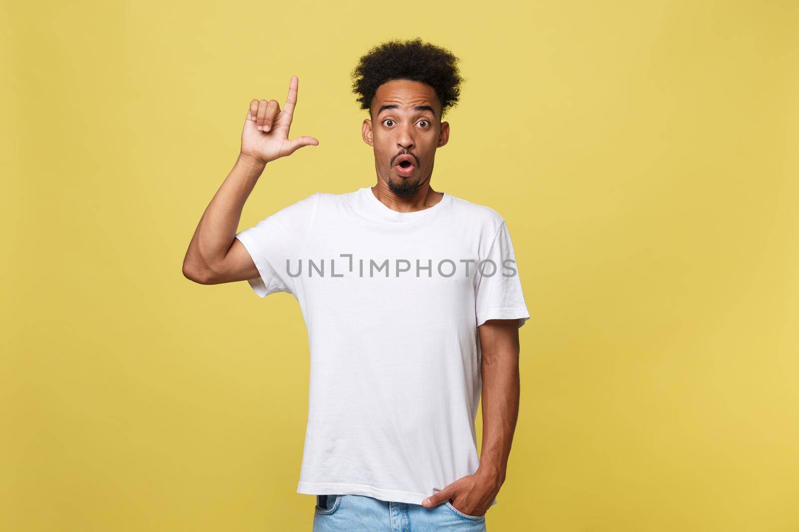 Astonished young African American man dressed in casual white shirt having excited fascinated look, pointing index finger at copy space on golden yellow background for your text or promotional content by Benzoix