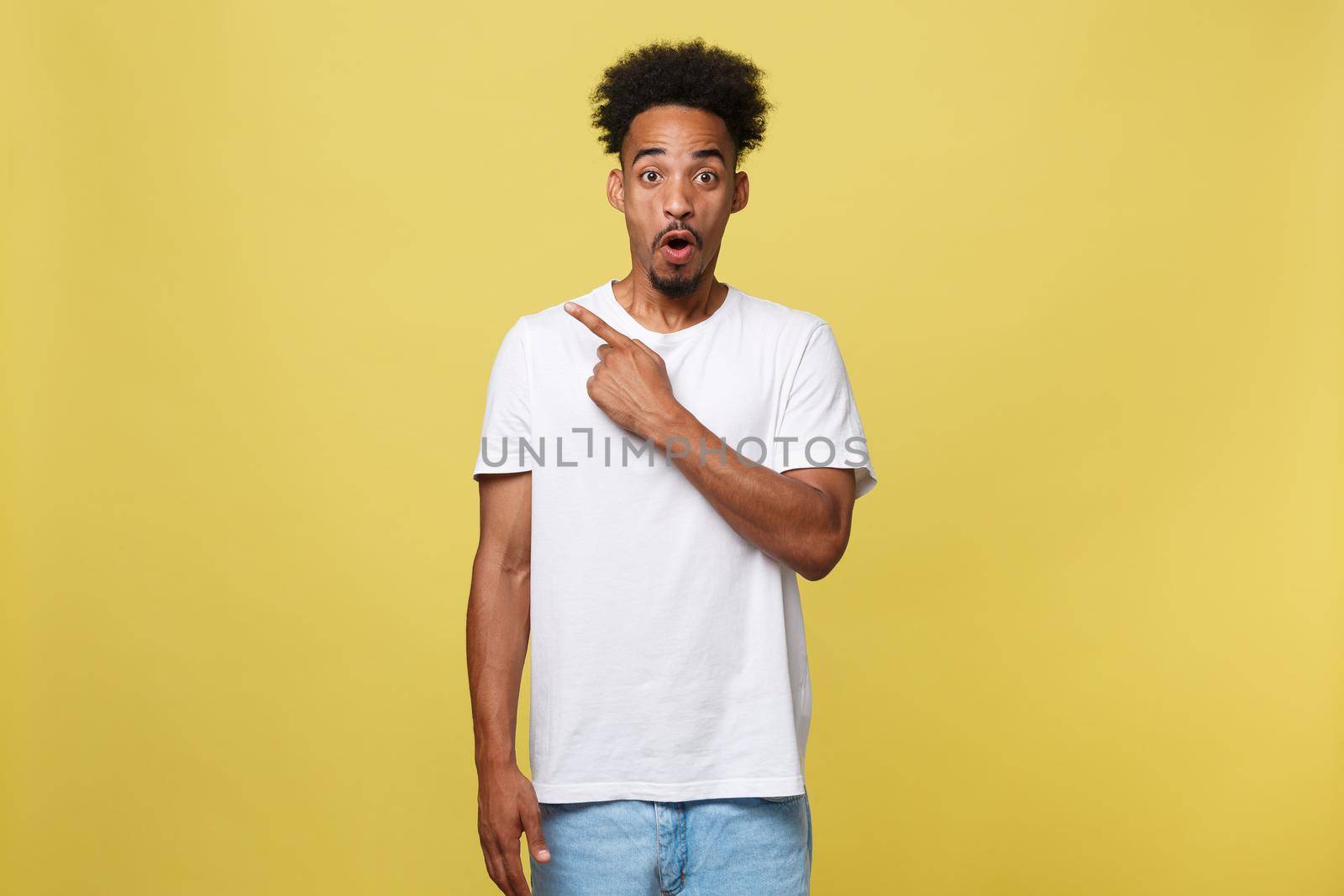 Astonished young African American man dressed in casual white shirt having excited fascinated look, pointing index finger at copy space on golden yellow background for your text or promotional content.