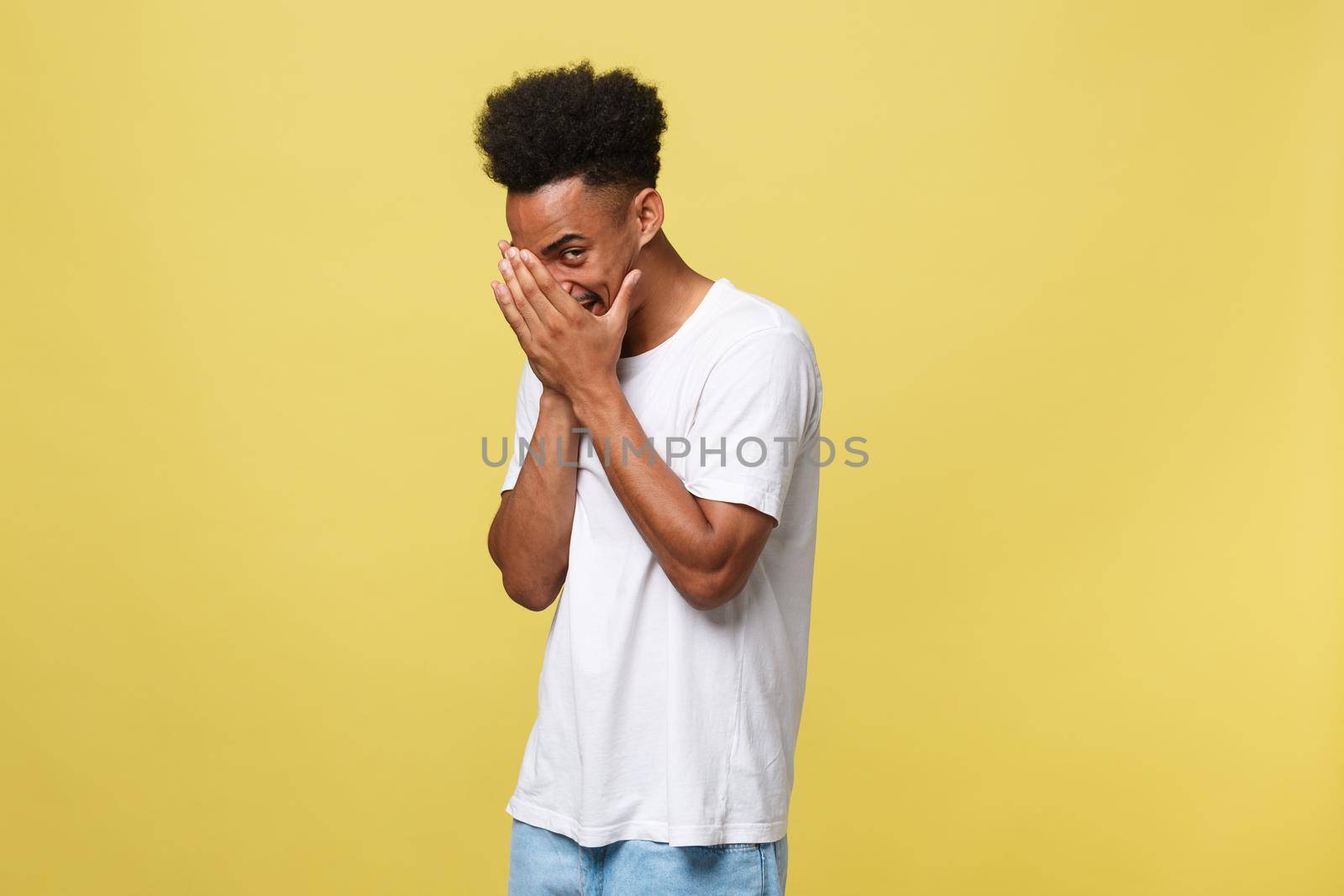 African american man with beard smiling having shy look peeking through fingers, covering face with hands looking confusedly broadly isolated over yellow background.
