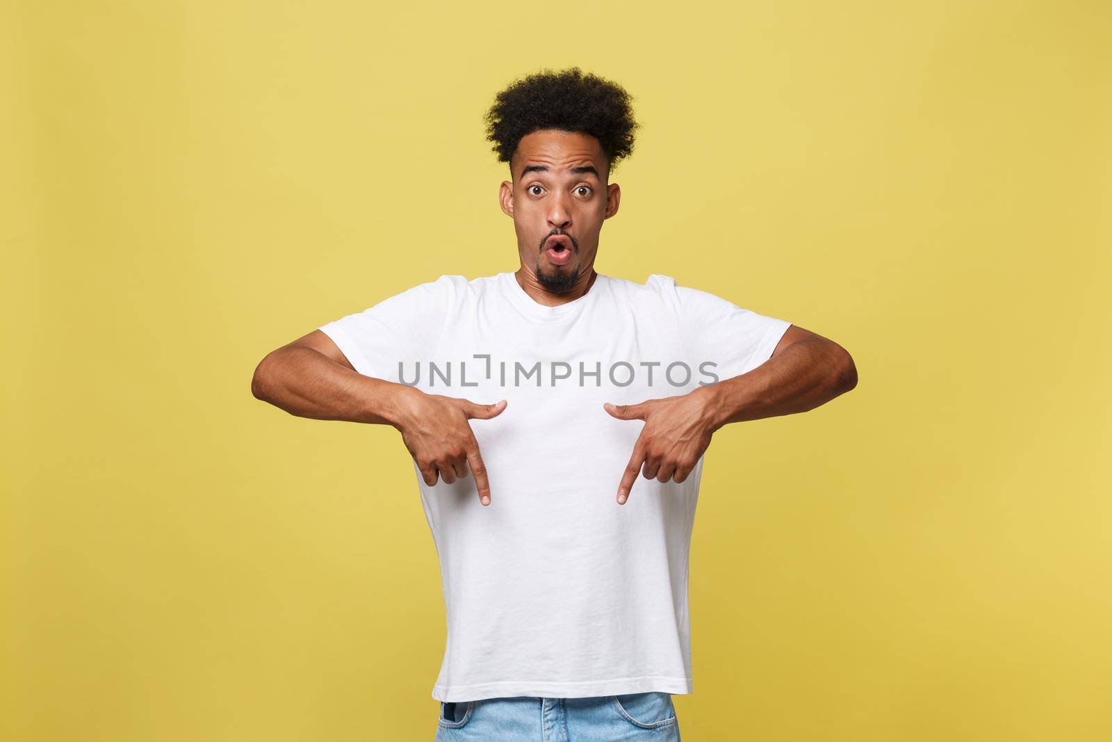 Astonished young African American man dressed in casual white shirt having excited fascinated look, pointing index finger at copy space on golden yellow background for your text or promotional content.