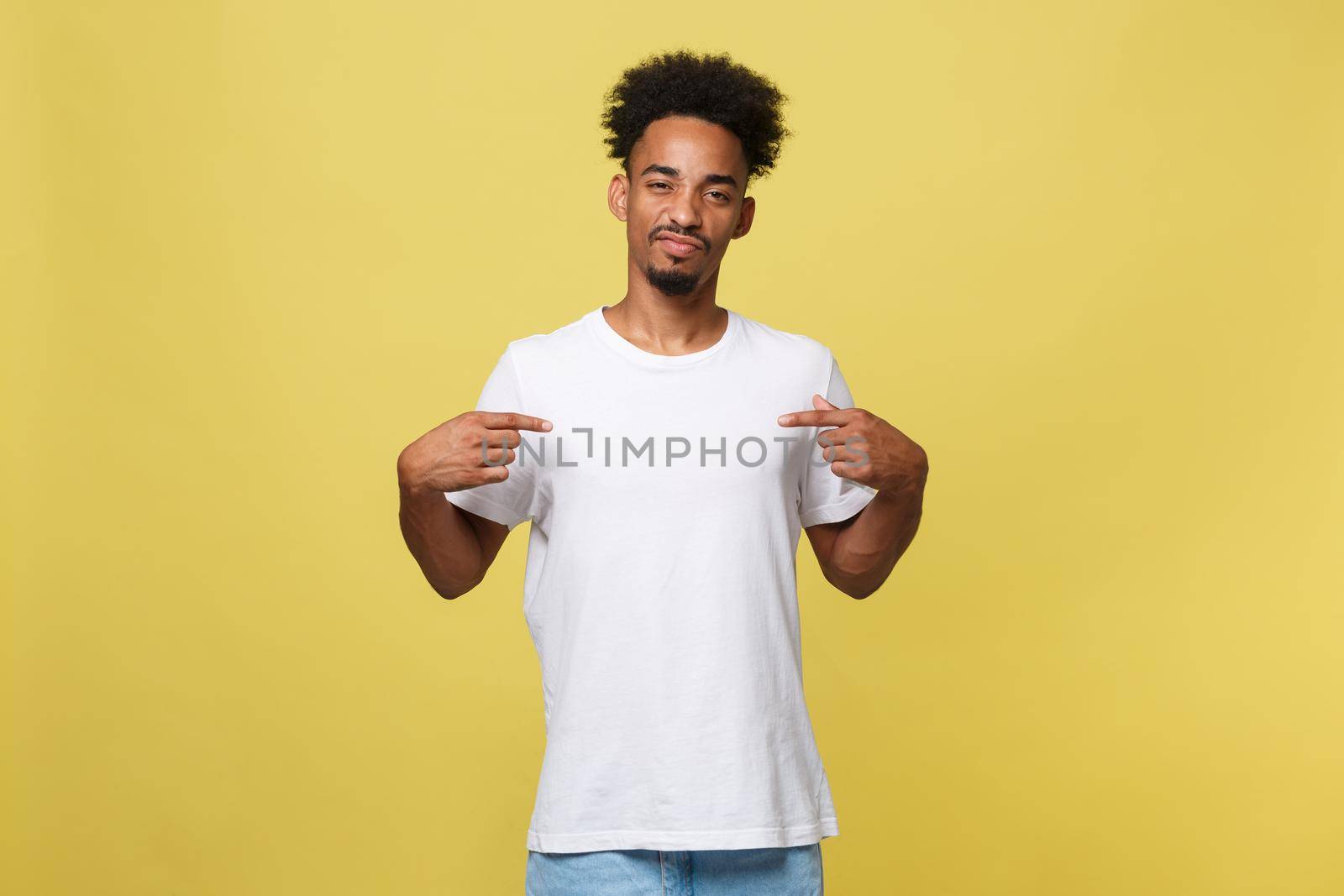 Portrait of handsome cheerful dark skinned male seller of clothes, dressed in casual white t shirt, indicates at blank copy space for your logo, isolated on yellow background. People, clothing, design.