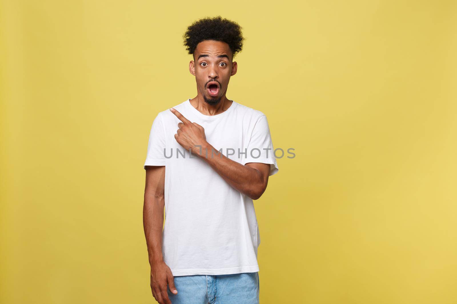 Astonished young African American man dressed in casual white shirt having excited fascinated look, pointing index finger at copy space on golden yellow background for your text or promotional content.