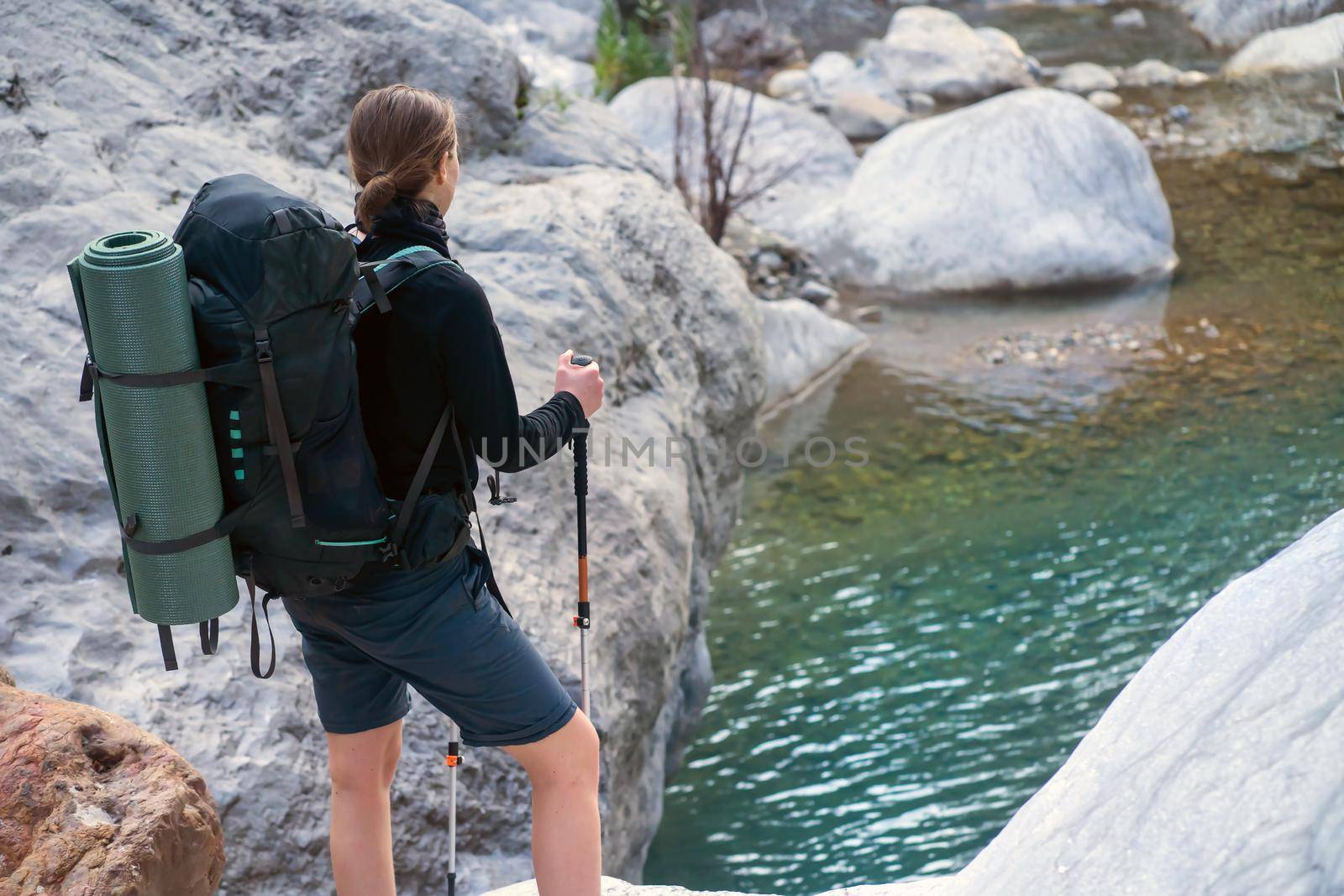 Young girl with a backpack and in hiking clothes holds trekking poles. Traveler looks at a transparent mountain river and rocks, is engaged in journey along the trail.