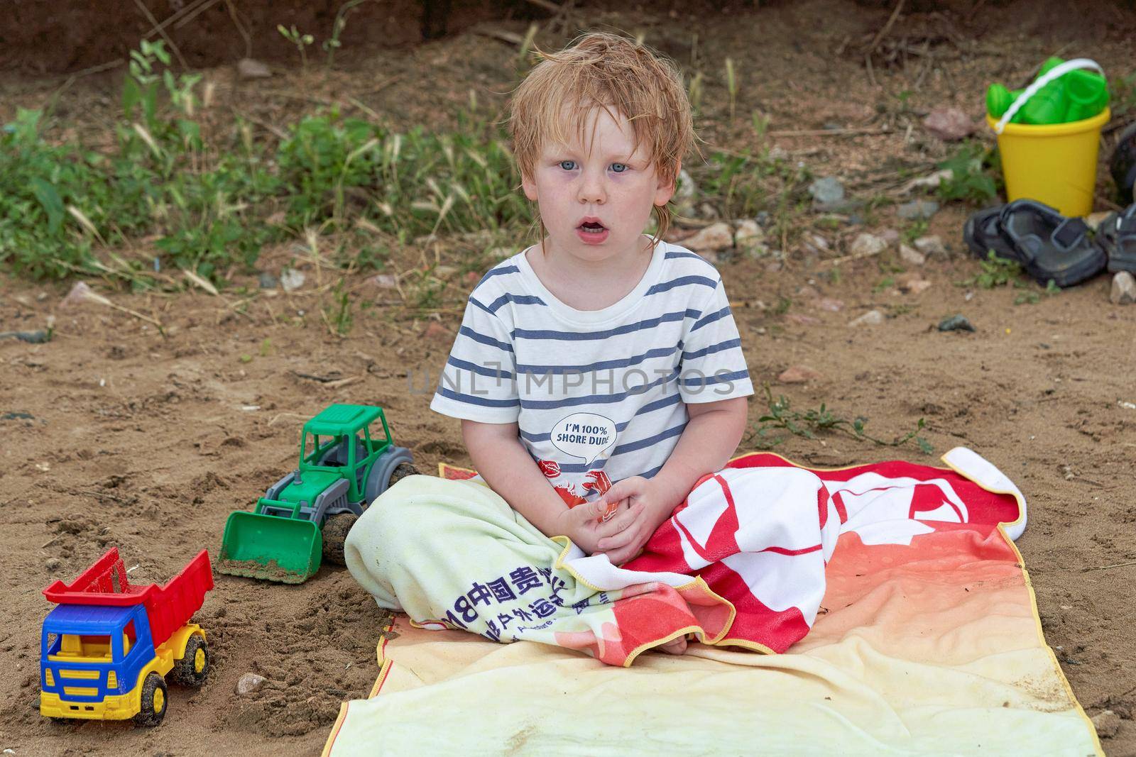 A little boy sits on a towel on a sandy beach among toys. Childrens holiday at the sea