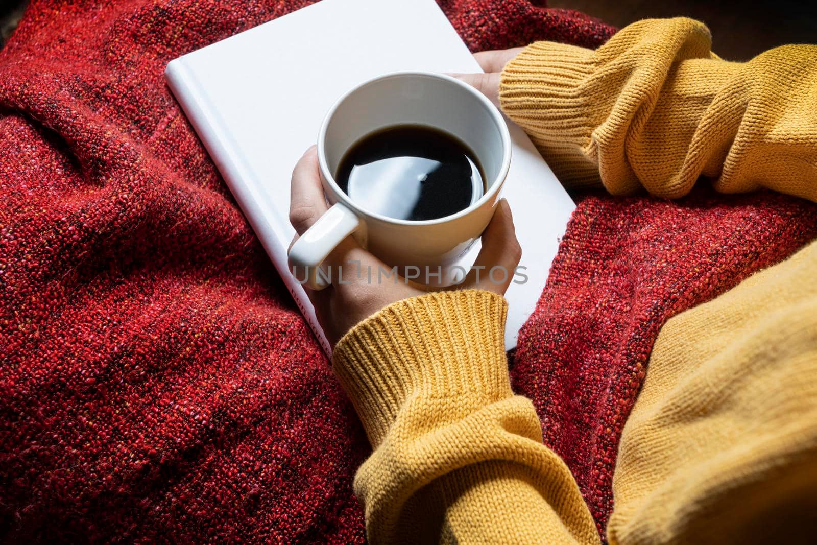 a woman with a red blanket on her legs relaxes by reading a book and enjoying a coffee