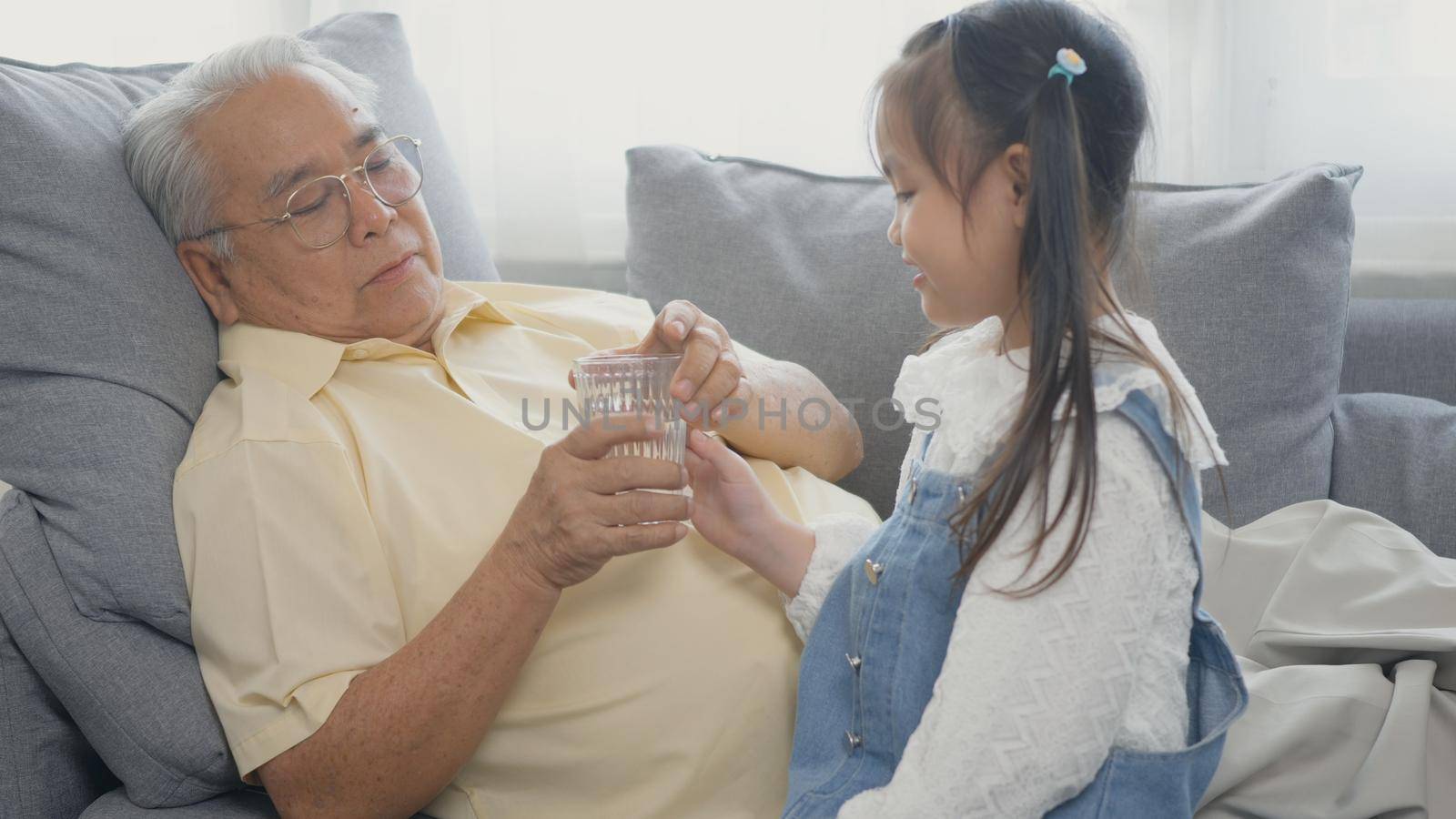 Asian granddaughter brought glass of water for grandpa to eat on the sofa in the living room. Happy little kid girl smiling giving wather to old man lie on sofa at home