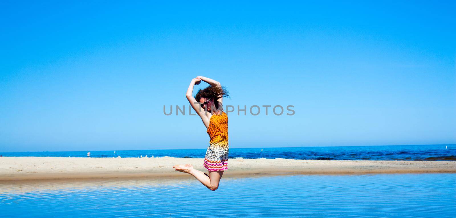 Woman with dark curly hair jumping in a bright blue lake with white sand dunes and a blue sky in the background by njproductions