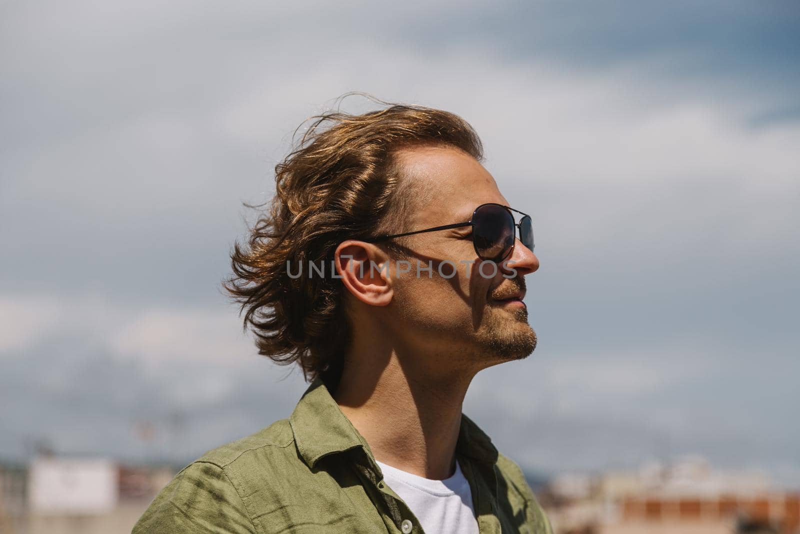 A happy man in sunglasses with long hair, standing outdoors on sunny day, smiling and looking to the side. by apavlin
