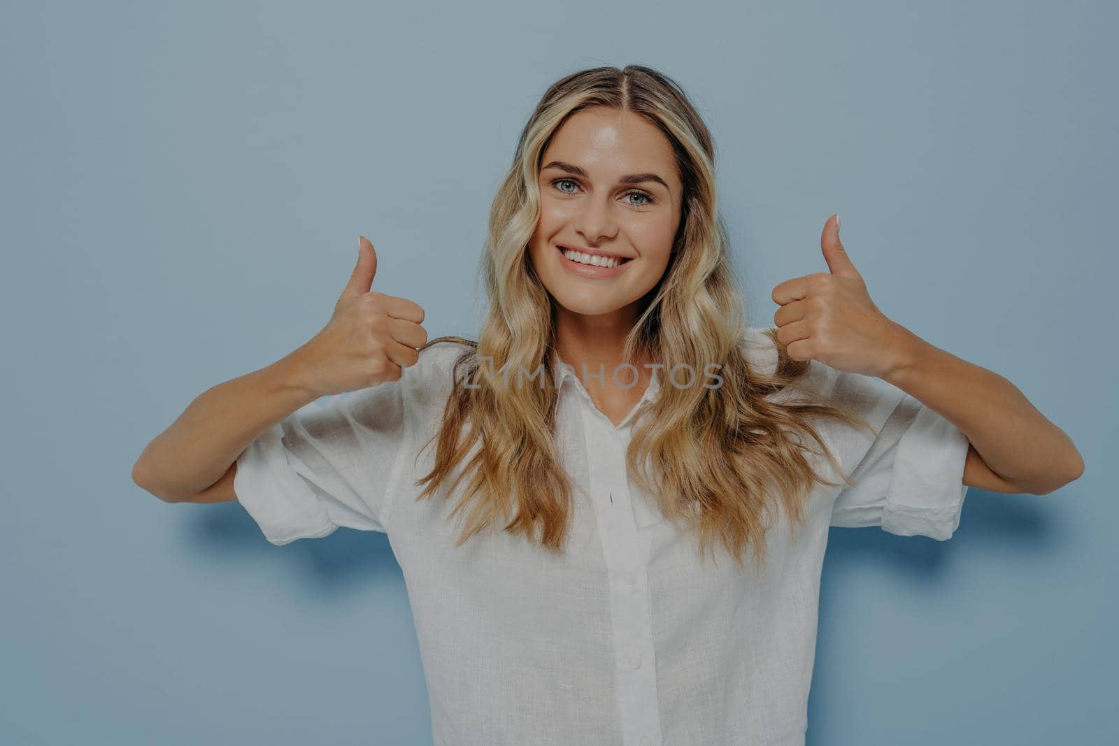 Blonde female dressed in white shirt expressing positive emotions while showing thumbs up gesture with both of her hands, displaying that she likes it, isolated next to blue wall