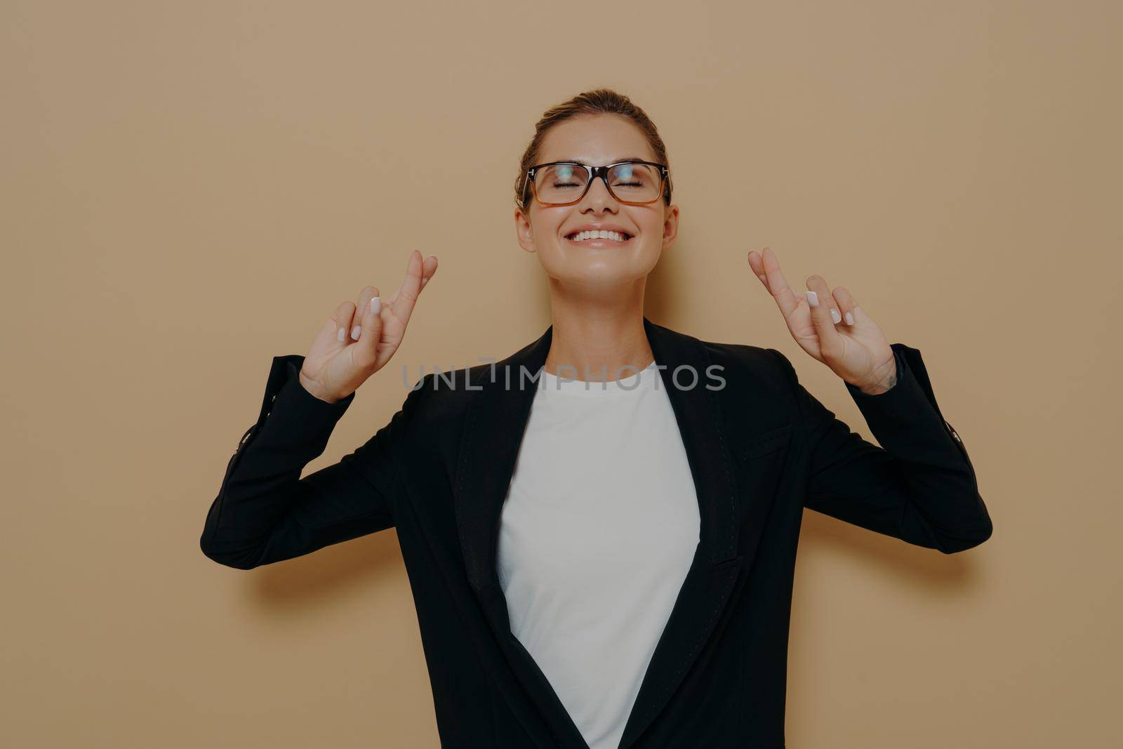 Young hopeful woman wearing eyeglasses in black blazer over white tshirt crossing her fingers with hope, keeping eyes closed and making wish while standing isolated over brown background