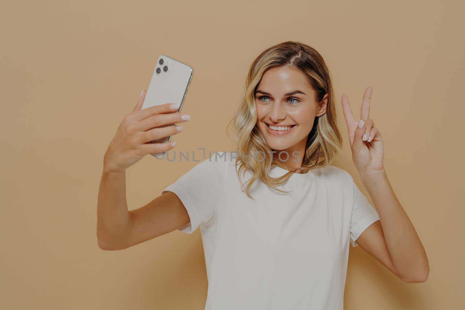 Cool joyful woman with blonde hair with smartphone in hand making selfie in studio against nude background and showing peace sign, taking picture of herself and smiling, dressed in white t shirt