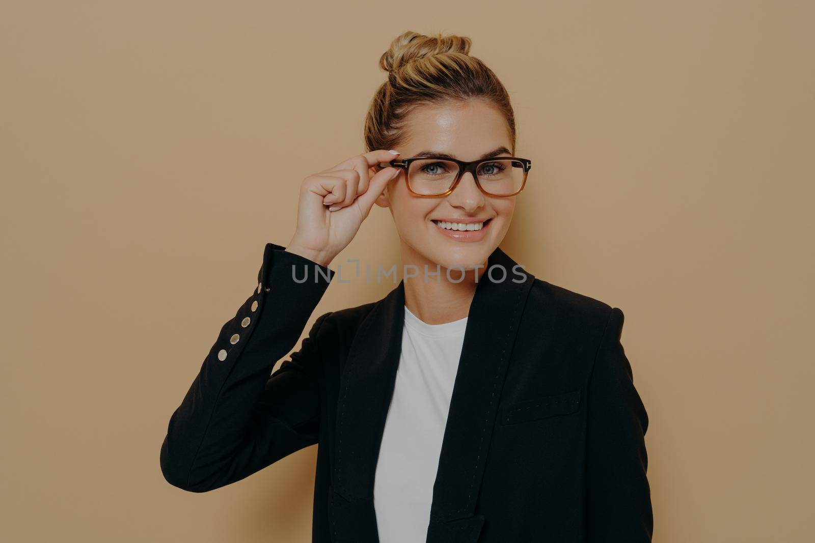 Portrait of happy satisfied female teacher in classic black suit smiling broadly and adjusting her glasses with hand while standing isolated over beige studio background with copy space