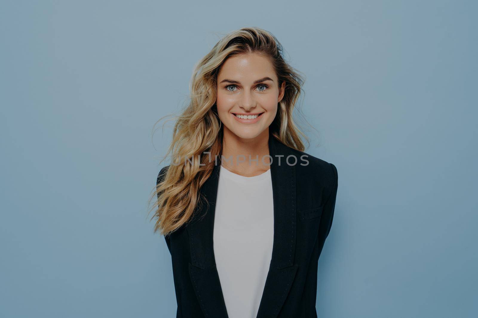 Vivacious blond woman in stylish blazer with happy beaming smile standing with hands behind her back by vkstock