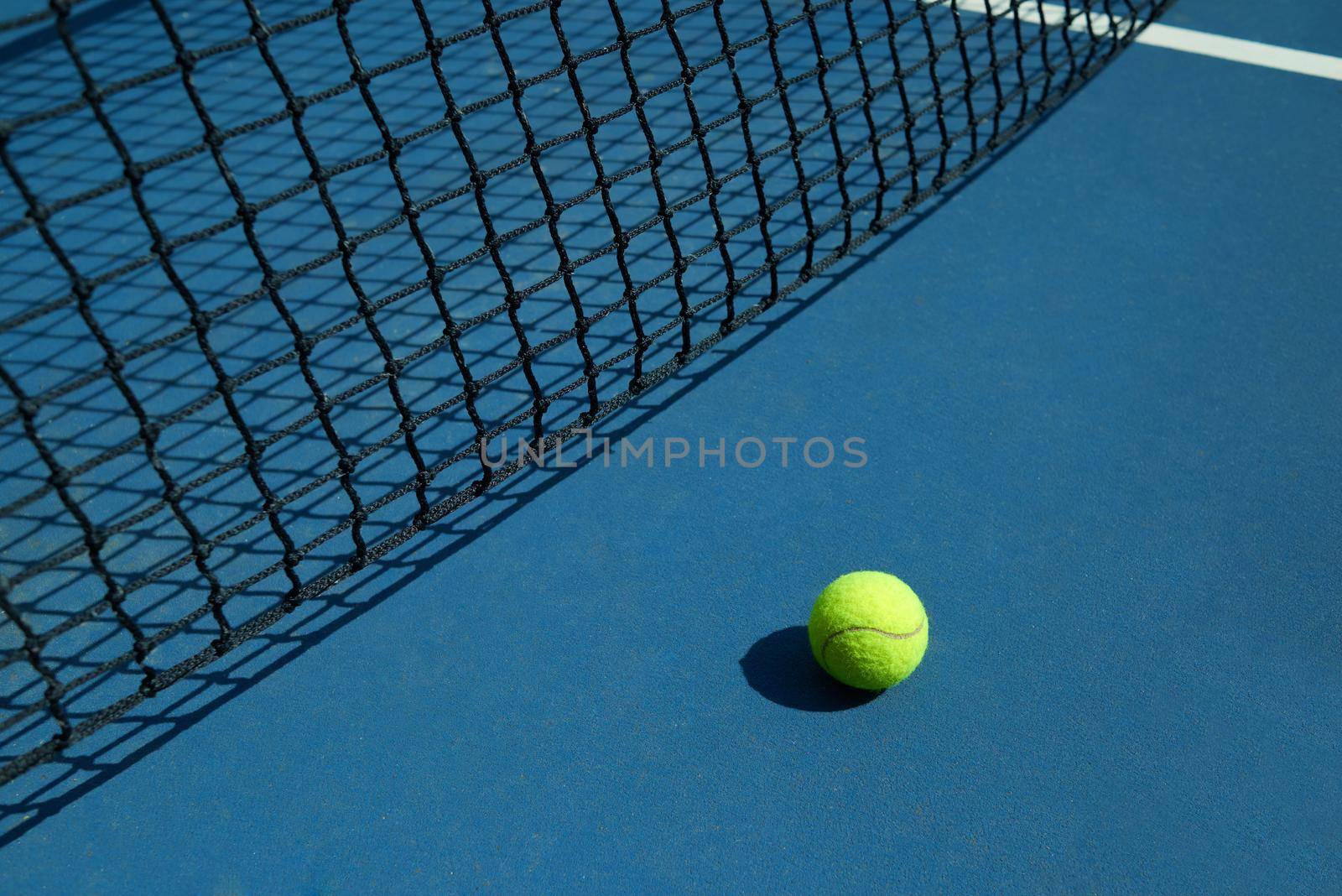 Yellow tennis ball is laying near black opened tennis court's net. Contrast image with satureted colors and shadows. Concept of tennis outfit photografing.