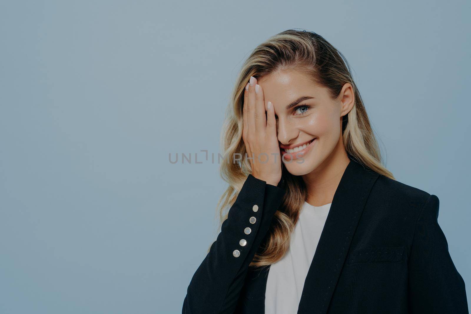 Blonde cute european woman in black coat covering half of her face with hand while smiling, expressing sadness with gesture, standing alone in front of blue background. Shy face expression concept