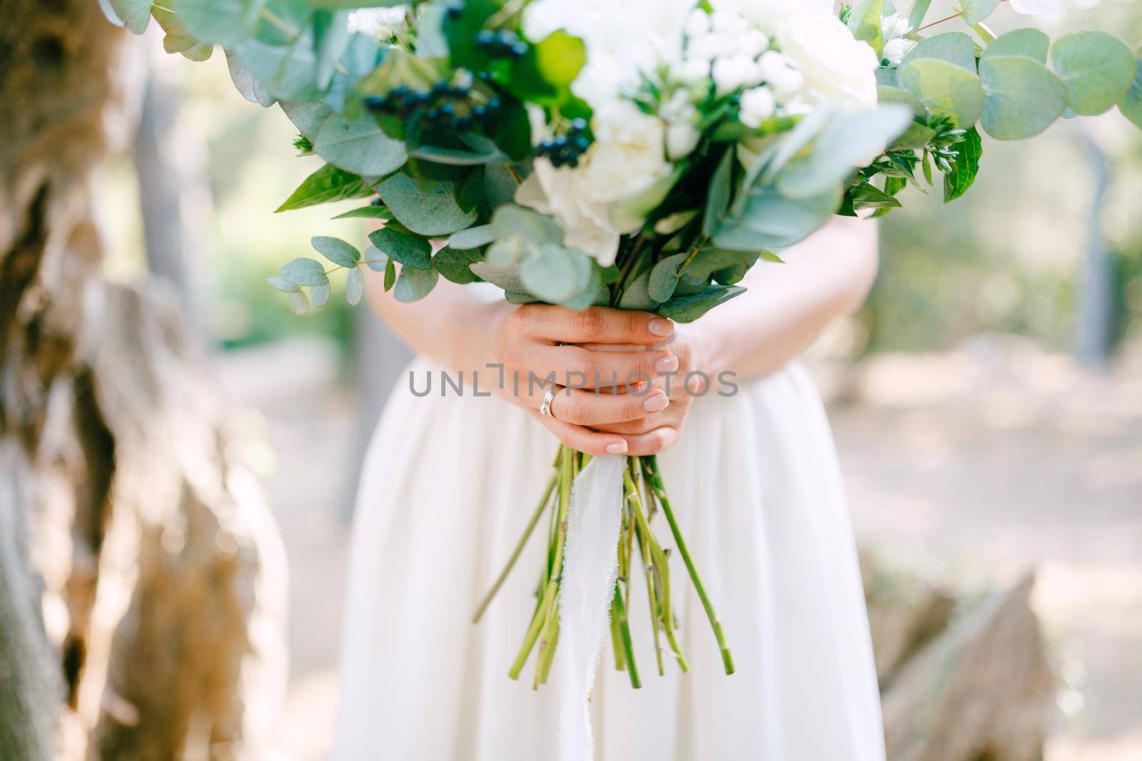 The bride is holding a wedding bouquet of roses, eucalyptus branches, delicate white flowers and dark berries in her hands, close-up by Nadtochiy