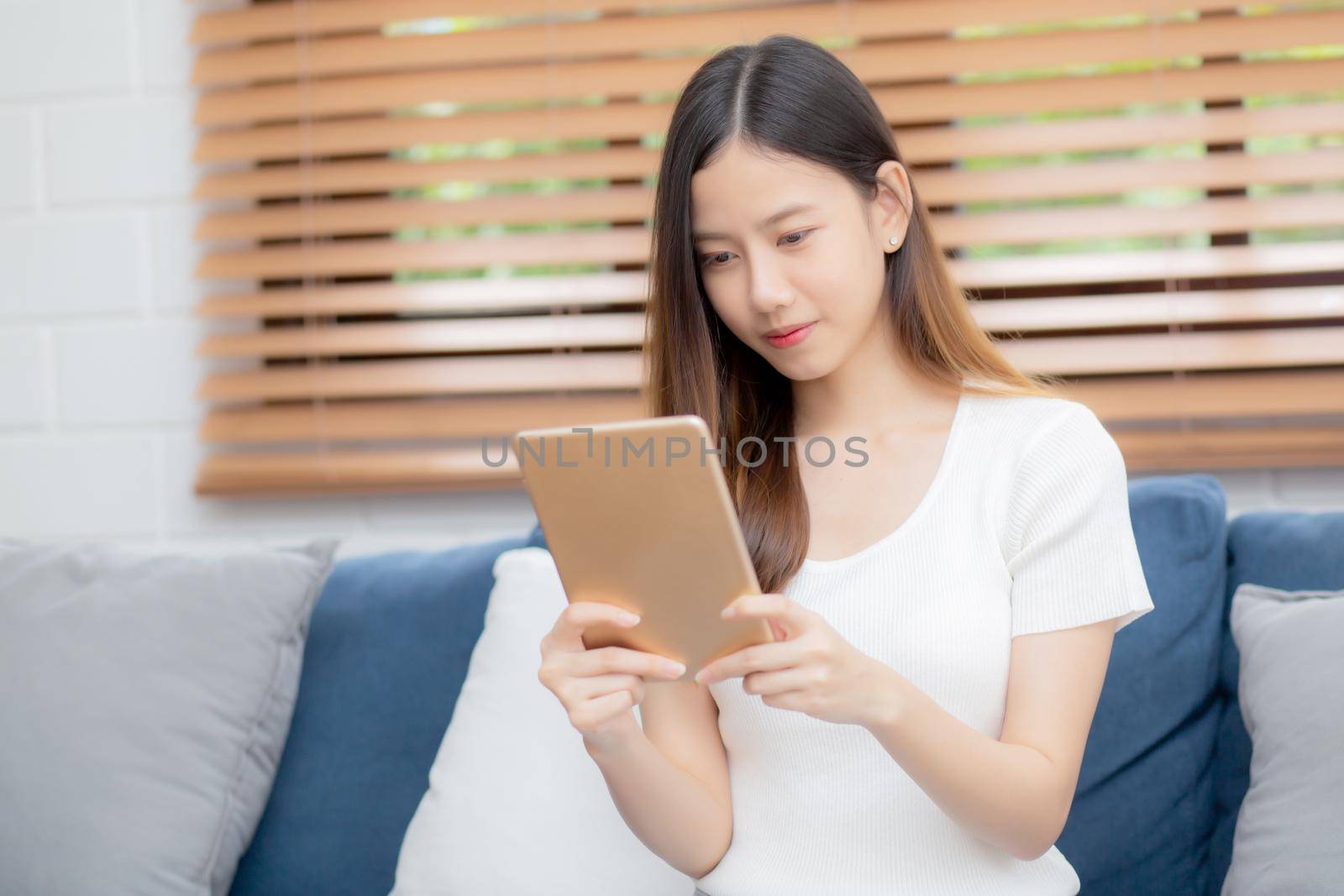 Young attractive asian woman resting using browsing tablet computer on sofa at home, happy girl sitting on couch relax reading digital gadget at house, communication and lifestyle concept.