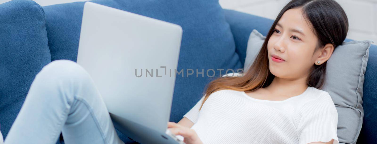 Young asian business woman smile and work from home with laptop computer online to internet on sofa in living room, freelance girl using notebook on couch with comfort, new normal, lifestyle concept.