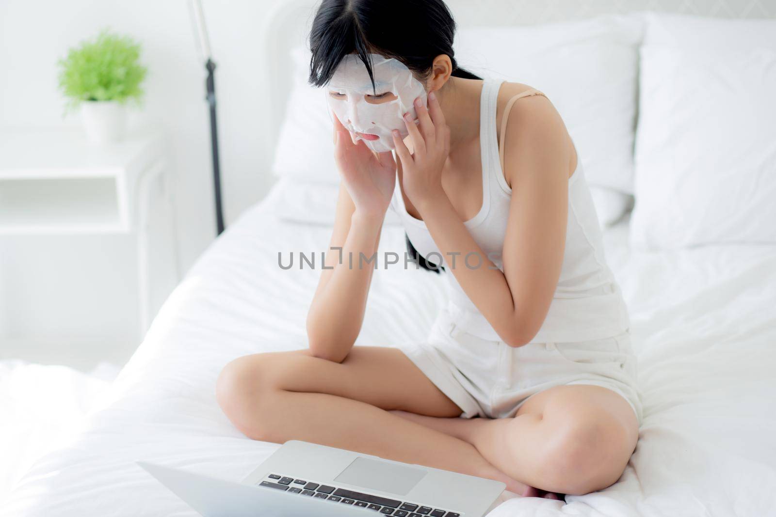 Beautiful young asian woman with sheet facial mask sitting working on laptop computer on bed at bedroom, beauty girl applying face mask for skincare of wrinkle at home, skin care, health and wellness. by nnudoo