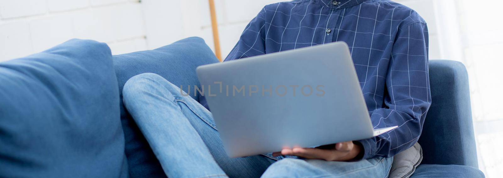 Young asian businessman work from home with laptop computer online to internet on sofa in living room, freelance man using notebook on couch with comfort, new normal, lifestyle concept. by nnudoo