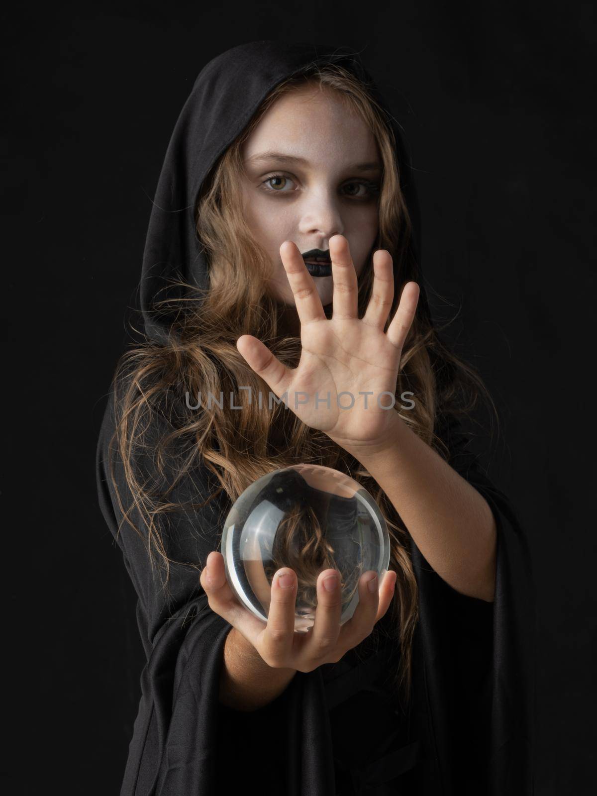 Little girl in Halloween witch costume and dark make-up holding crystal ball isolated on black background