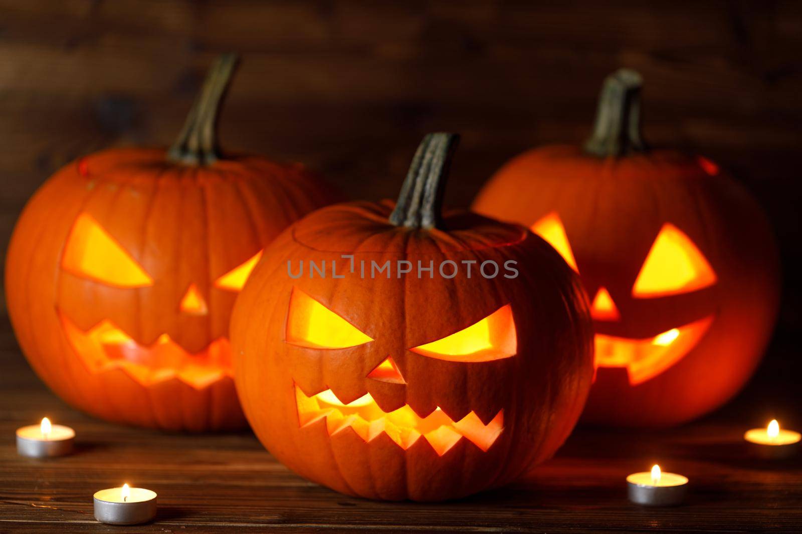 Three glowing Halloween Pumpkin lanterns and burning candles on dark wooden background