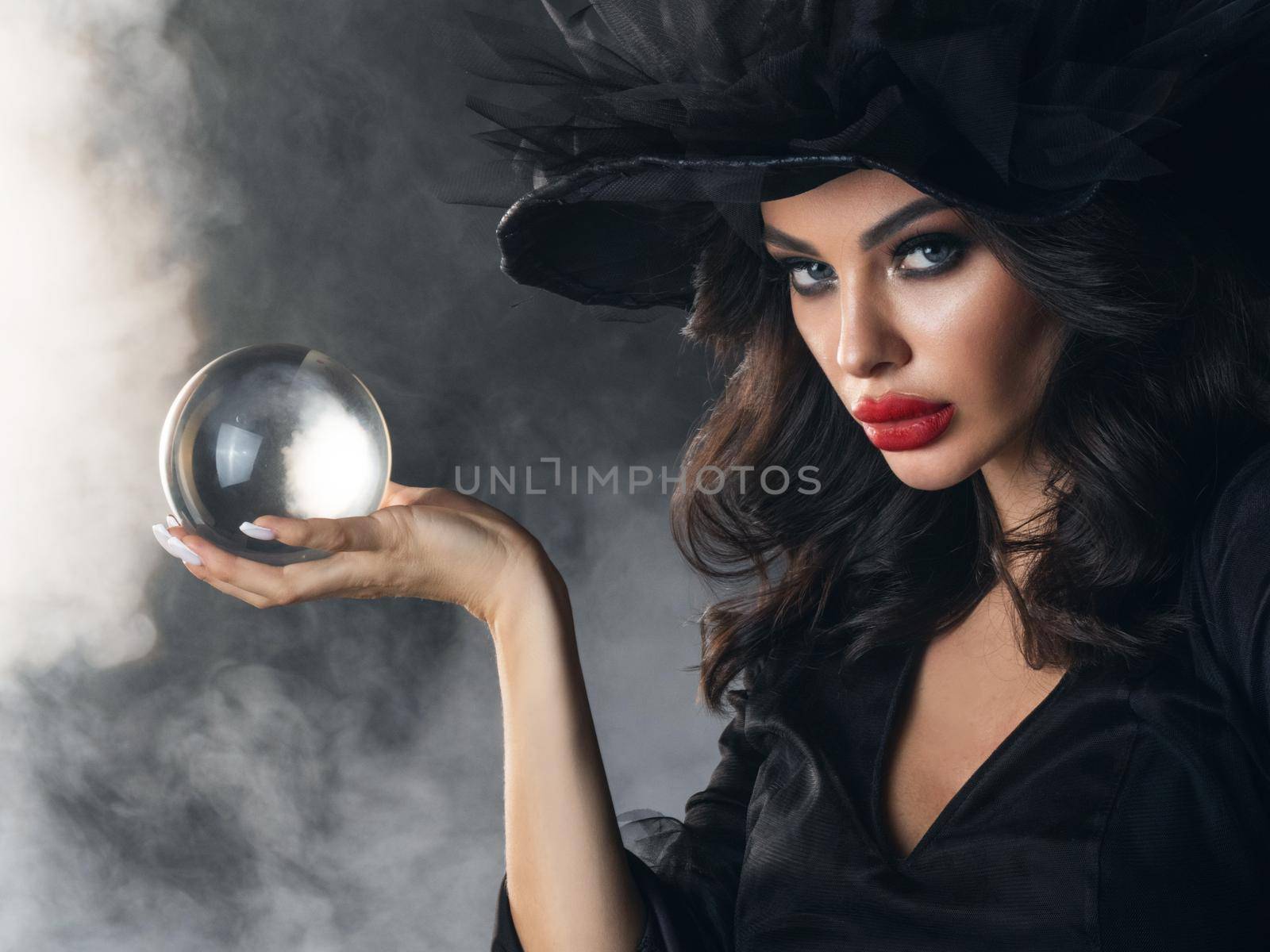 Woman in Halloween witch costume future teller showing crystal ball. Beautiful sexy model girl in sinister costume and make up over foggy background