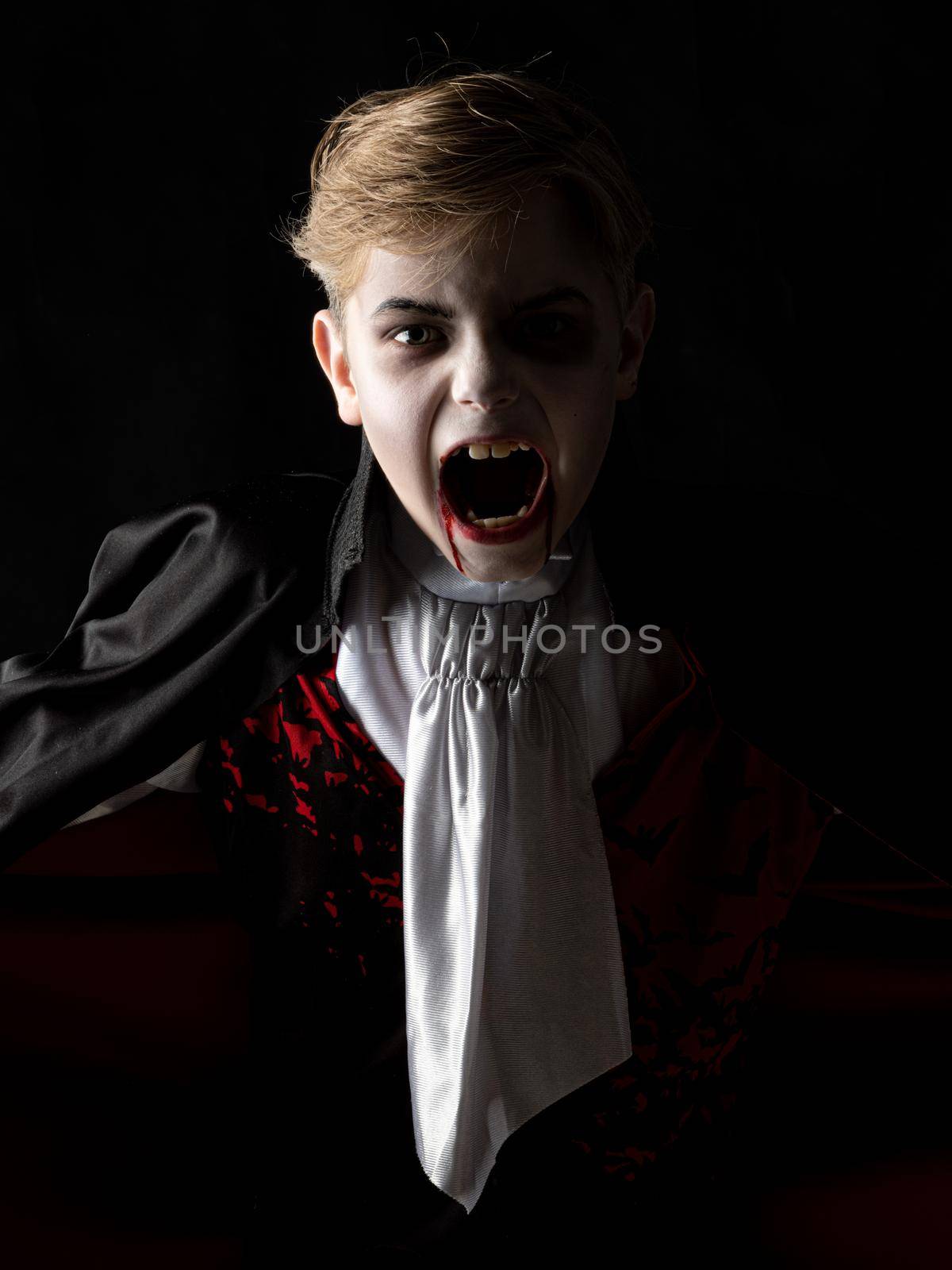 Portrait of boy wearing Halloween vampire makeup and costume cloak bare his teeth on black background.
