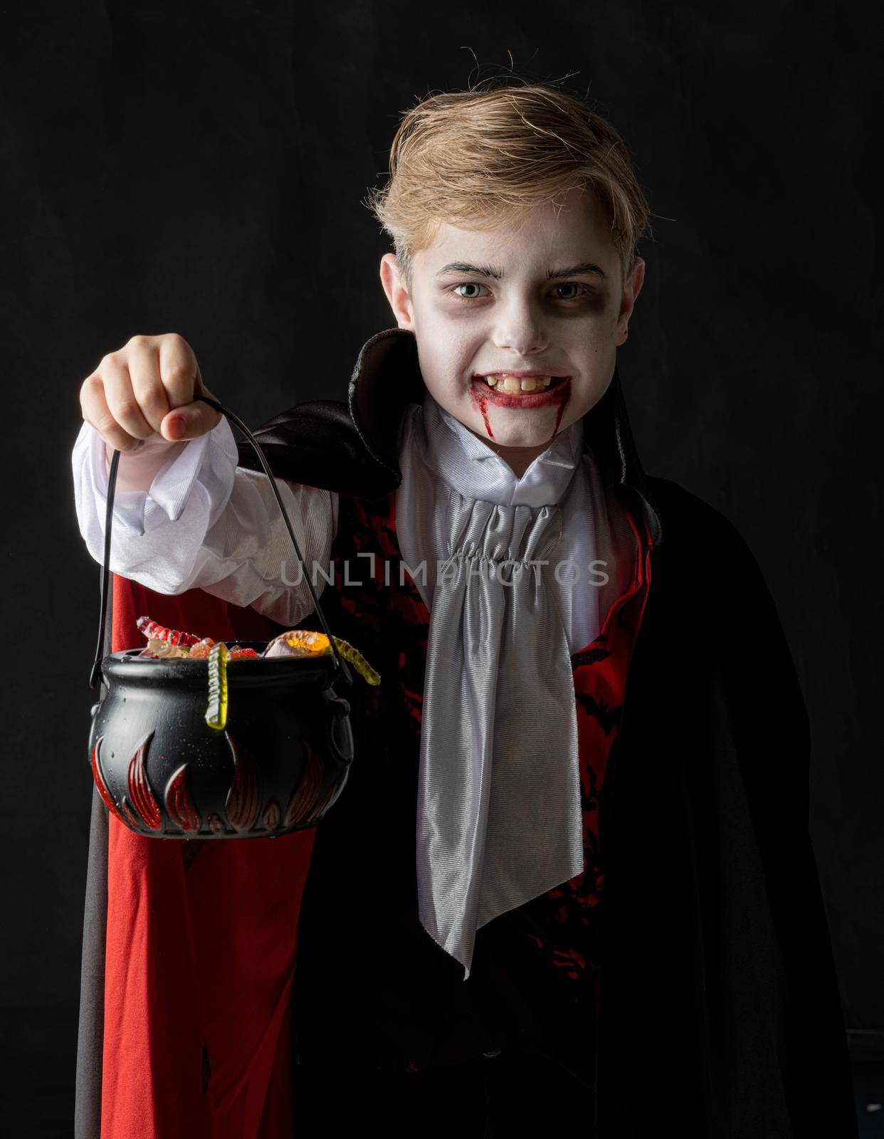 Boy with cauldron basket and worm sweets dressed as vampire for Halloween party going trick or treating. Studio portrait isolated over black background