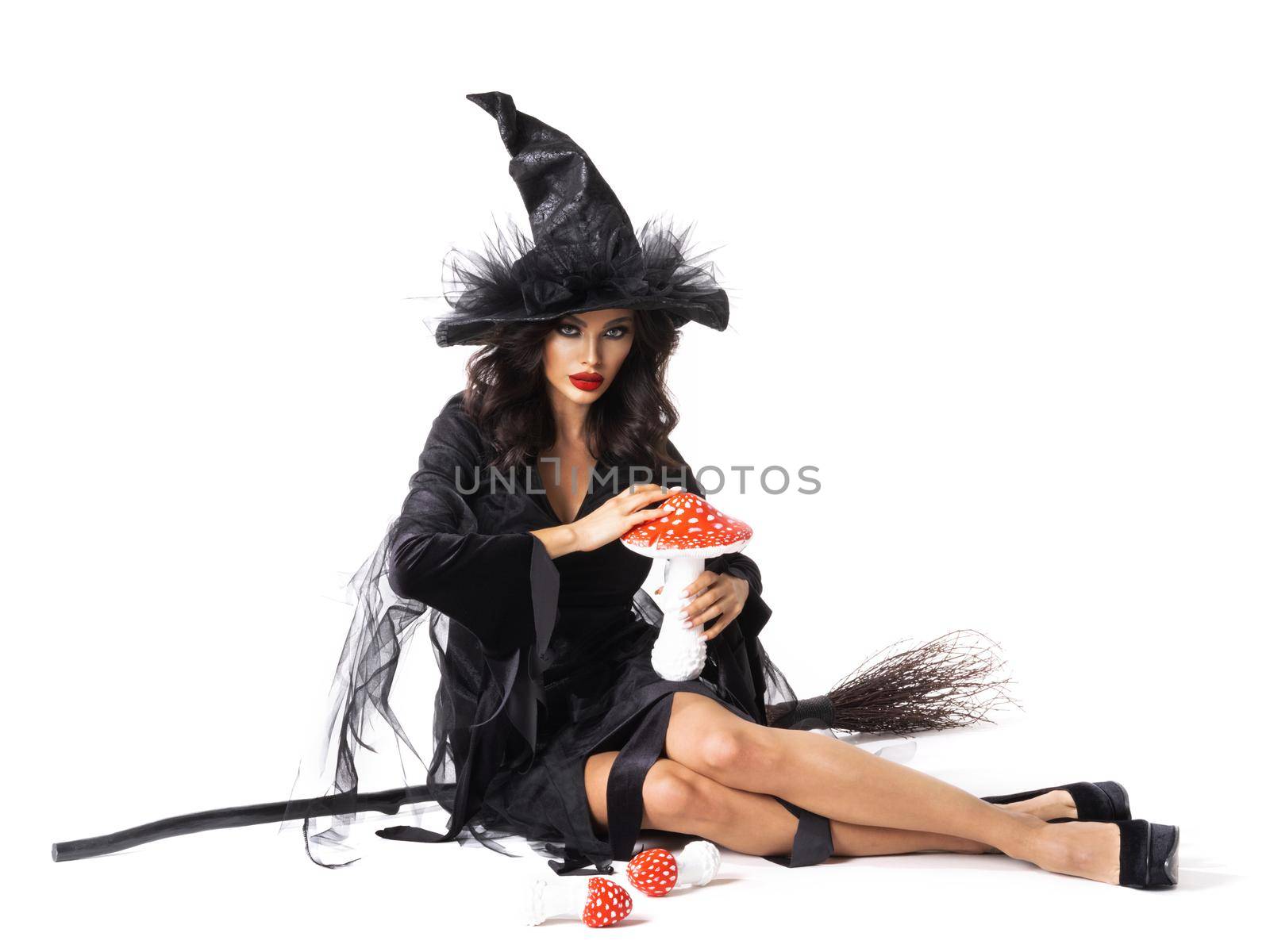 Halloween witch with broom and fly agarics poisonous mushrooms sitting isolated on white background, beauty woman portrait in party costume