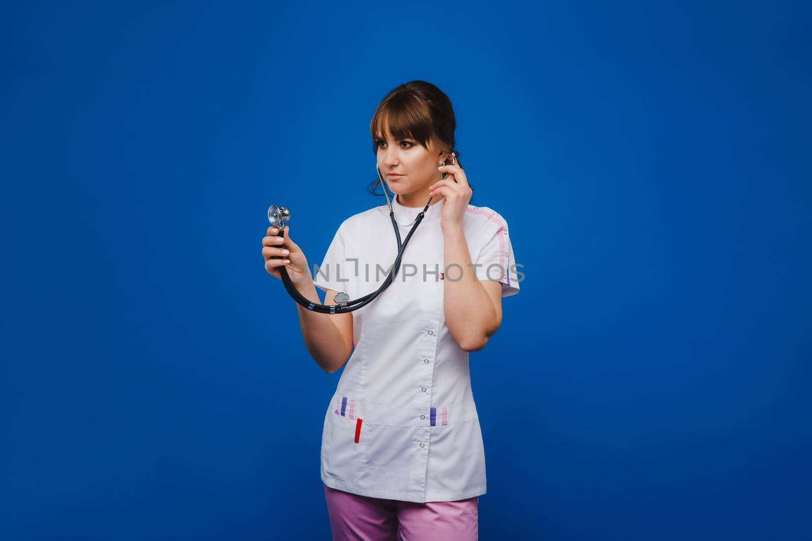 A female doctor, gesticulating, checks the heartbeat in the doctor's office at the hospital with a stethoscope isolated on a blue background by Lobachad