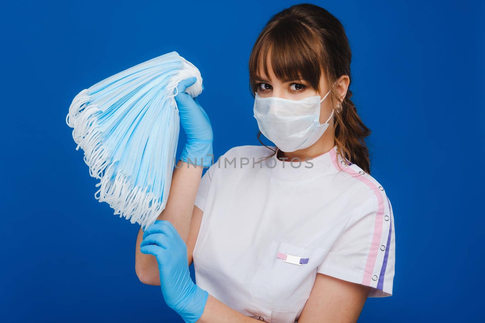 A young girl doctor in a medical mask holds a lot of masks in her hands. A nurse poses on a blue background by Lobachad