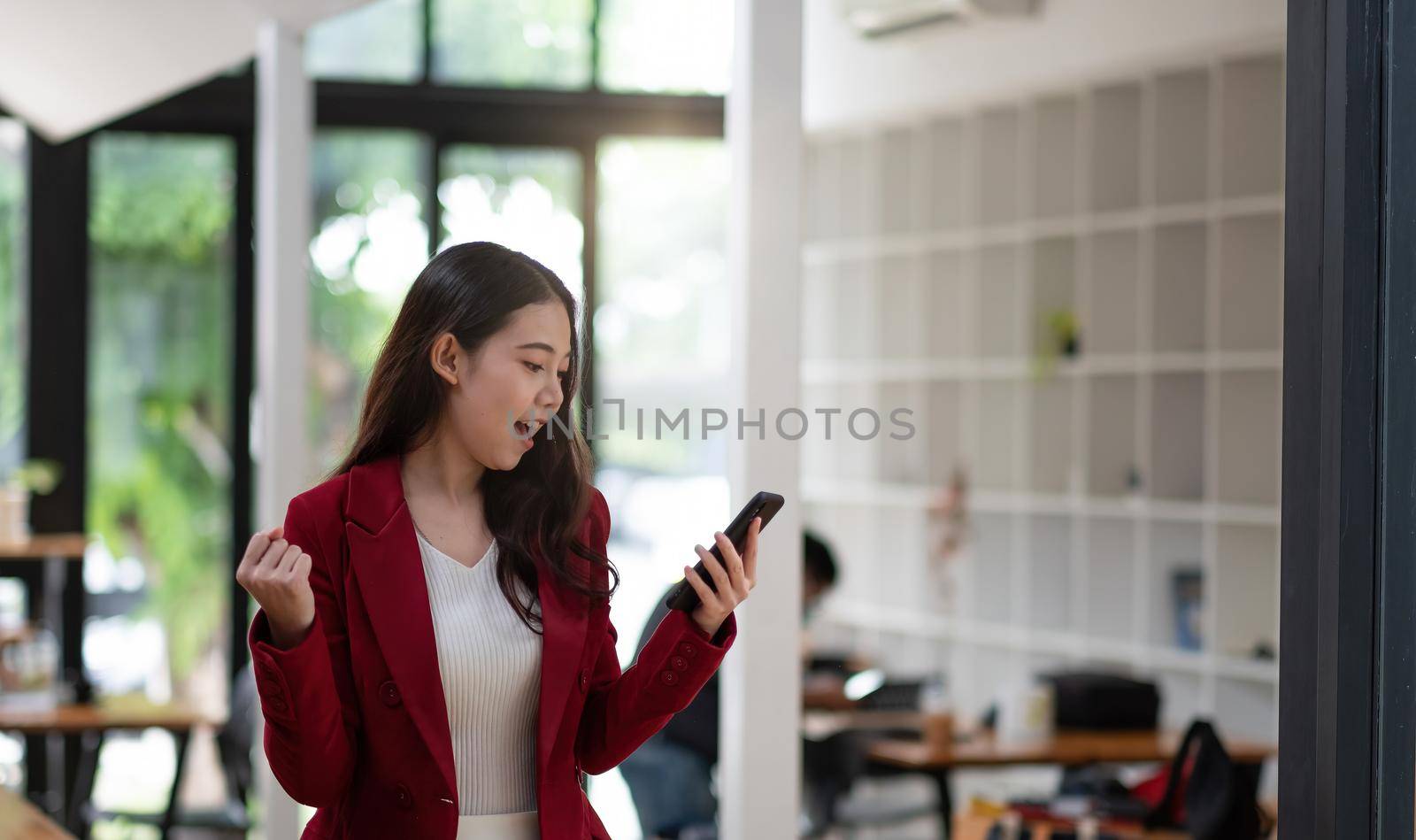 Smiling Asian woman holding mobile phone with fist hand and excited for success in office by nateemee