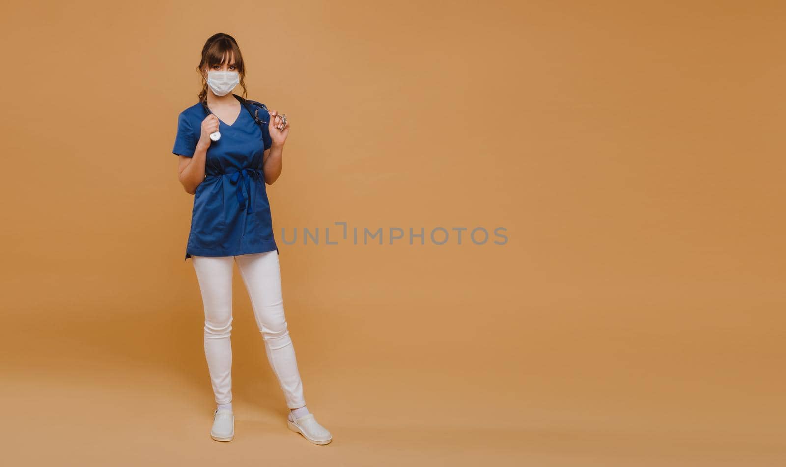 A girl doctor stands in a medical mask, isolated on a brown background.