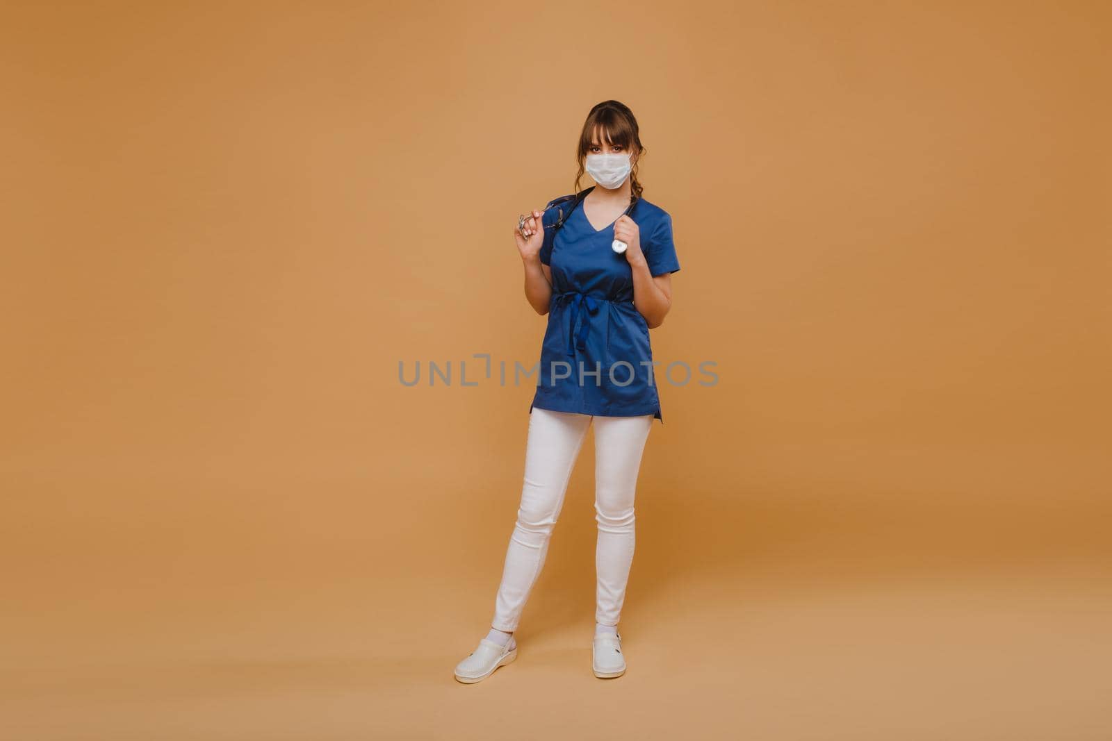 A girl doctor stands in a medical mask, isolated on a brown background.