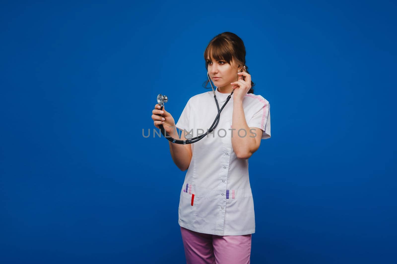 A female doctor, gesticulating, checks the heartbeat in the doctor's office at the hospital with a stethoscope isolated on a blue background by Lobachad