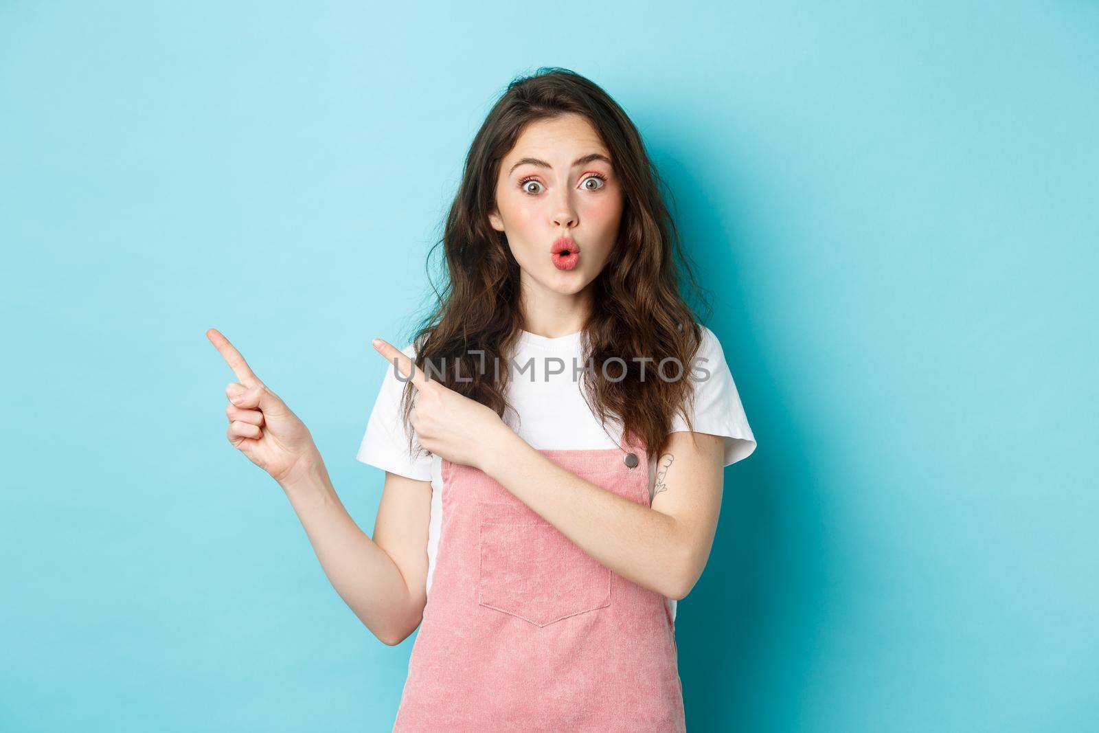 Wow check this out. Amazed brunette female model pucker lips and look intrigued, pointing fingers left at something interesting, showing banner, blue background.