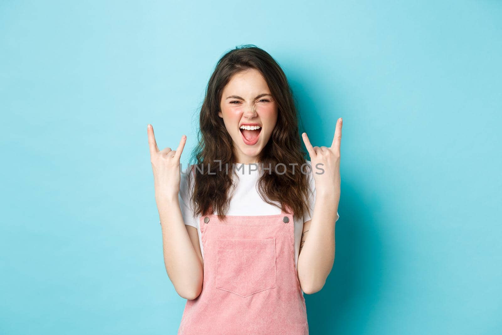 Portrait of excited woman enjoying concert or awesome event, showing rock n roll horns gesture and shouting with joy, having fun, standing over blue background.