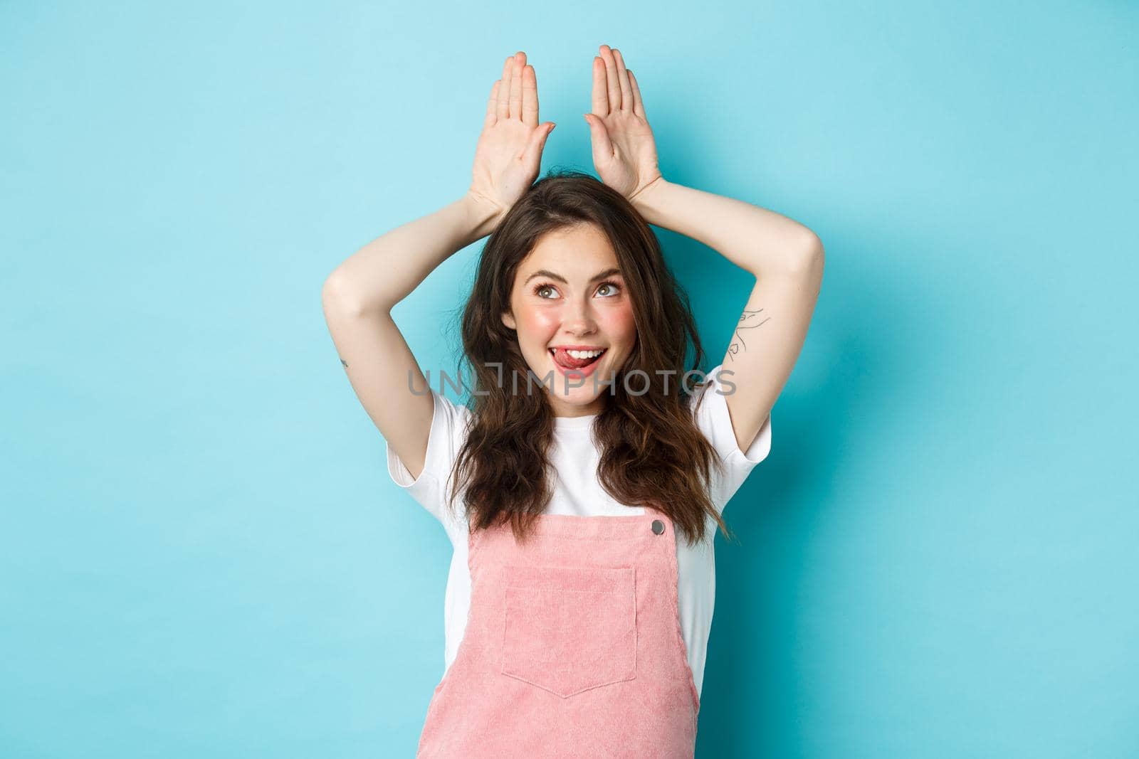 Image of happy young woman celebrating Easter, showing bunny ears with hands on head and smiling cheerful, enjoying spring holiday, standing over blue background by Benzoix