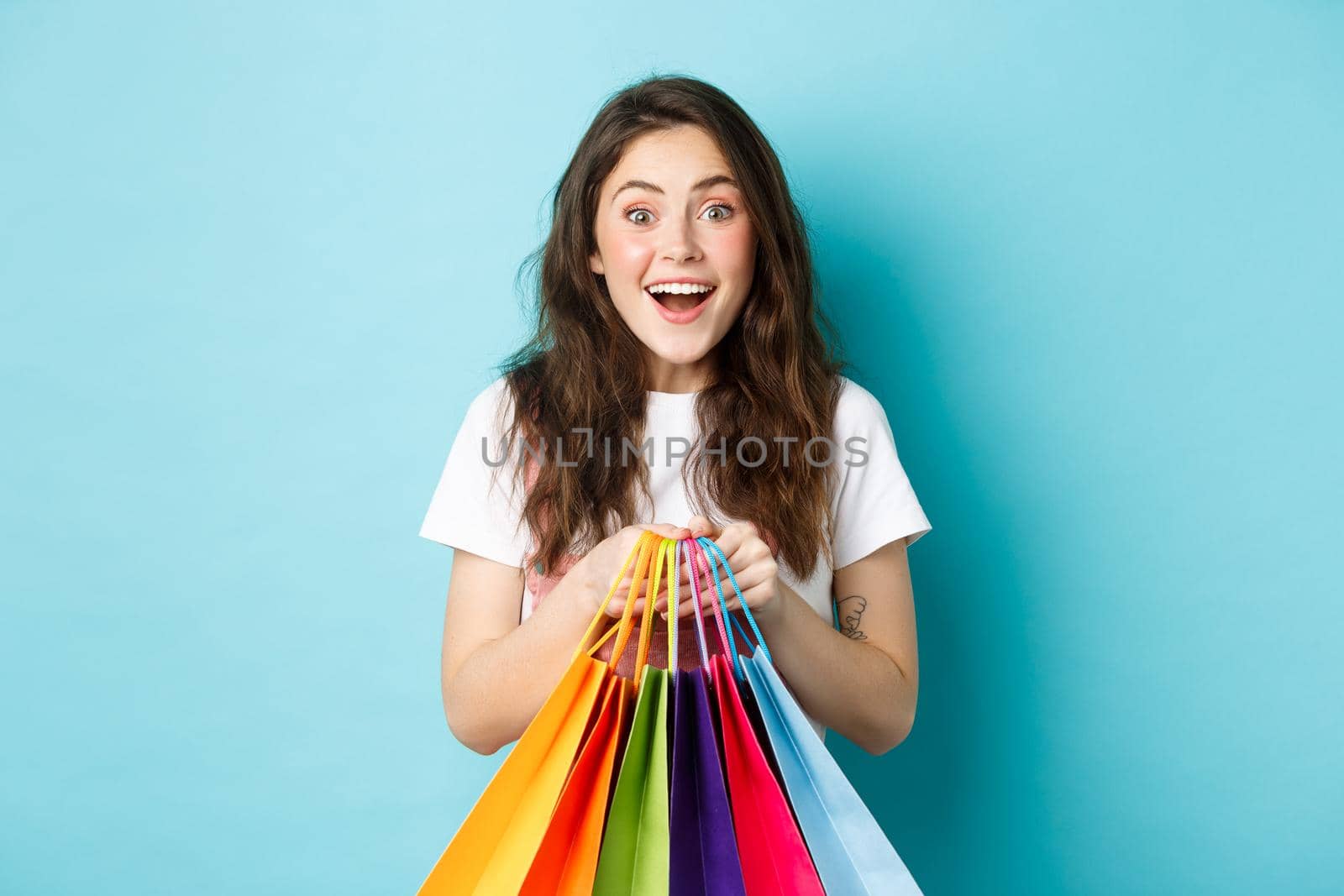 Portrait of excited girl shopper, holding shopping bags, buying in stores and smiling amazed, enjoy discounts, standing over blue background.