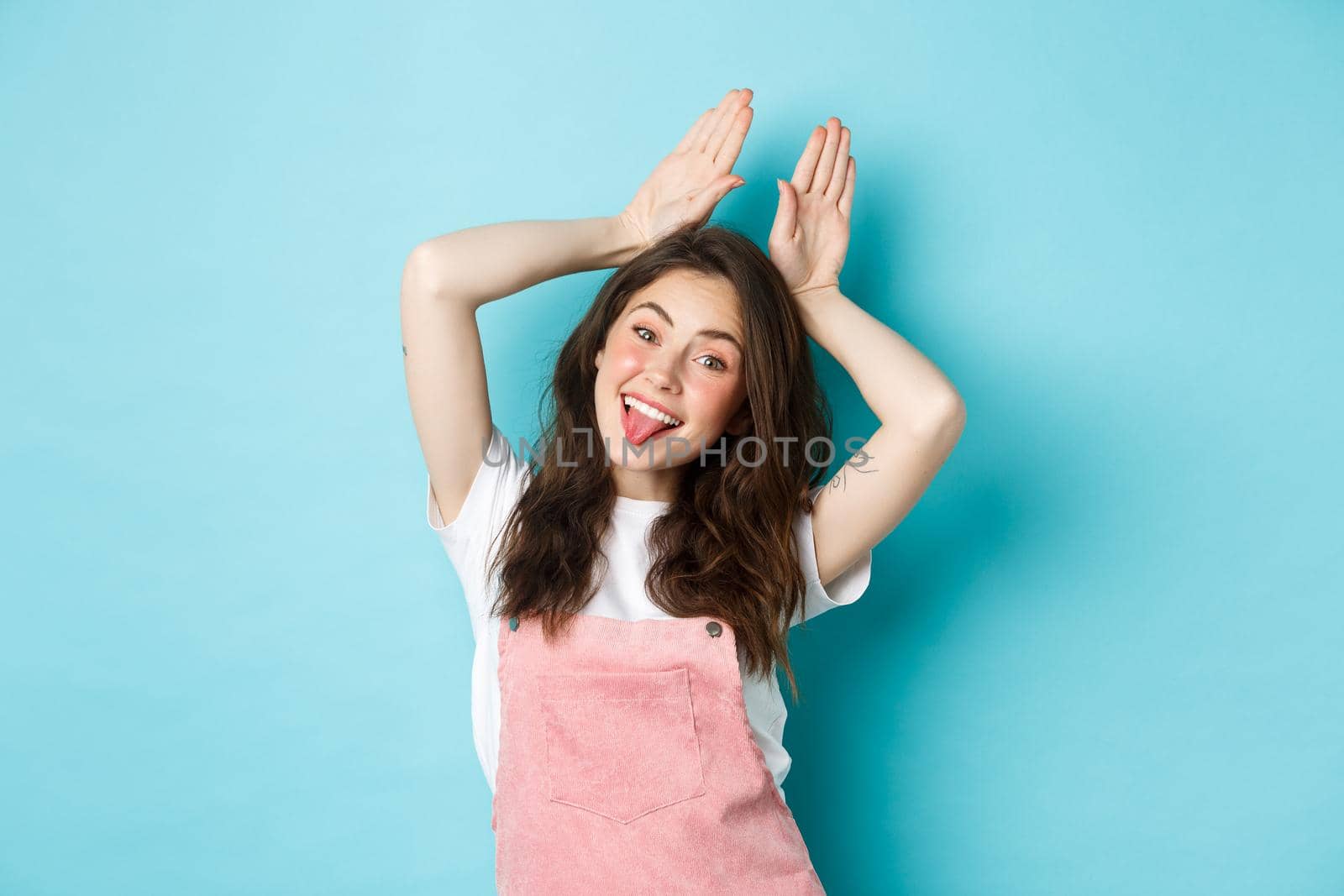 Portrait of beautiful glamour girl celebrating Easter, showing bunny rabbit ears with hands above head, smiling joyful, standing over blue background.