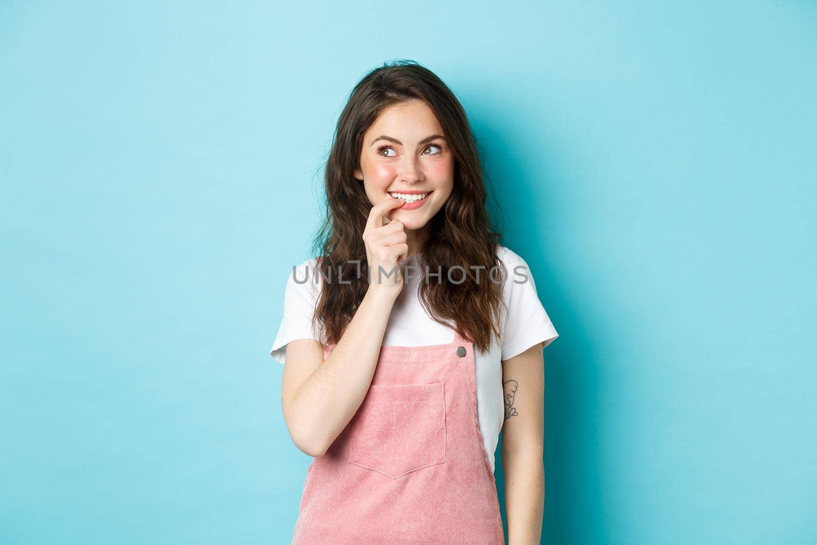 Thoughtful silly girl with cute curly hairstyle, biting finger and looking left at promo logo, smiling thinking about tasting ot trying something, standing over blue background.