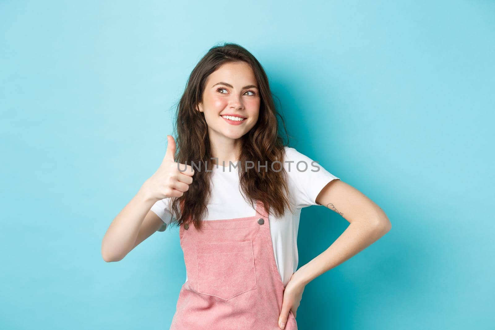 Portrait of smiling cute girl with curly hairstyle, showing thumb up and looking thoughtful at upper left corner banner, praising store sale, standing against blue background by Benzoix