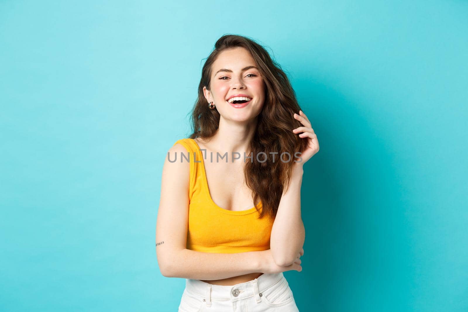 Summer and lifestyle concept. Portrait of attractive woman laughing and playing with her hair, standing in yellow cropped top, going on holiday, standing against blue background.