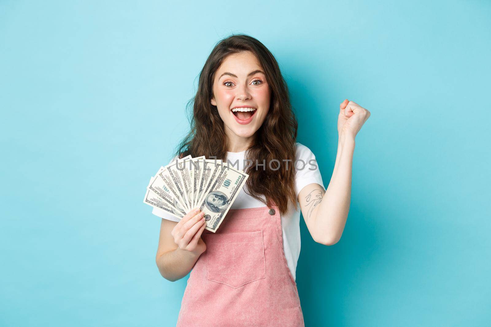 Excited smiling girl fist pump and hold money prize, winning cash, receive income from something, standing happy against blue background.