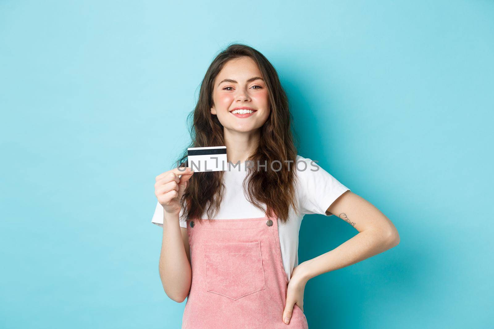 Proud smiling girl looking boastful, showing her plastic credit card, going to buy something, shopping contactless, standing over blue background.