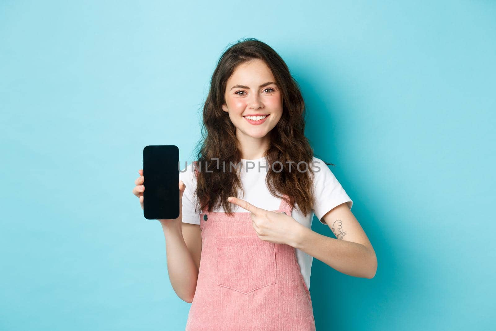 Happy smiling brunette girl pointing finger at empty smartphone screen, recommending online shop or mobile app, standing against blue background.